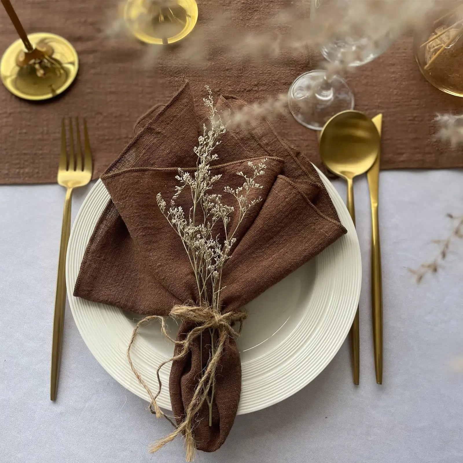 Une table de salle à manger élégante, avec une assiette blanche, des couverts dorés et un serviette de table marron ornée de fleurs blanches, créant une atmosphère chaleureuse et raffinée.