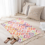 Tapis décoratif multicolore avec motifs géométriques, coussins ornés et pantoufles en osier sur le sol d'une pièce aux rideaux blancs.