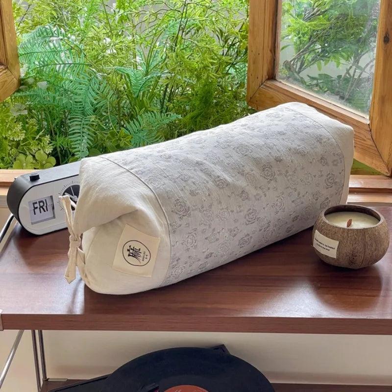 Un coussin cylindrique de style scandinave est posé sur une étagère en bois devant une fenêtre avec une vue verdoyante, accompagné d'un réveil et d'une bougie dans un récipient en bois.