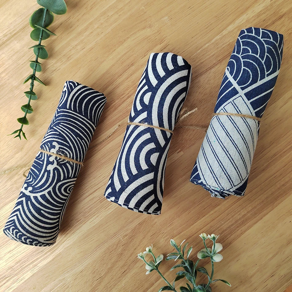 Des formes géométriques et des motifs rayés bleus et blancs sur des cylindres en tissu, accompagnés de plantes vertes, créent une composition graphique et harmonieuse sur une surface en bois.