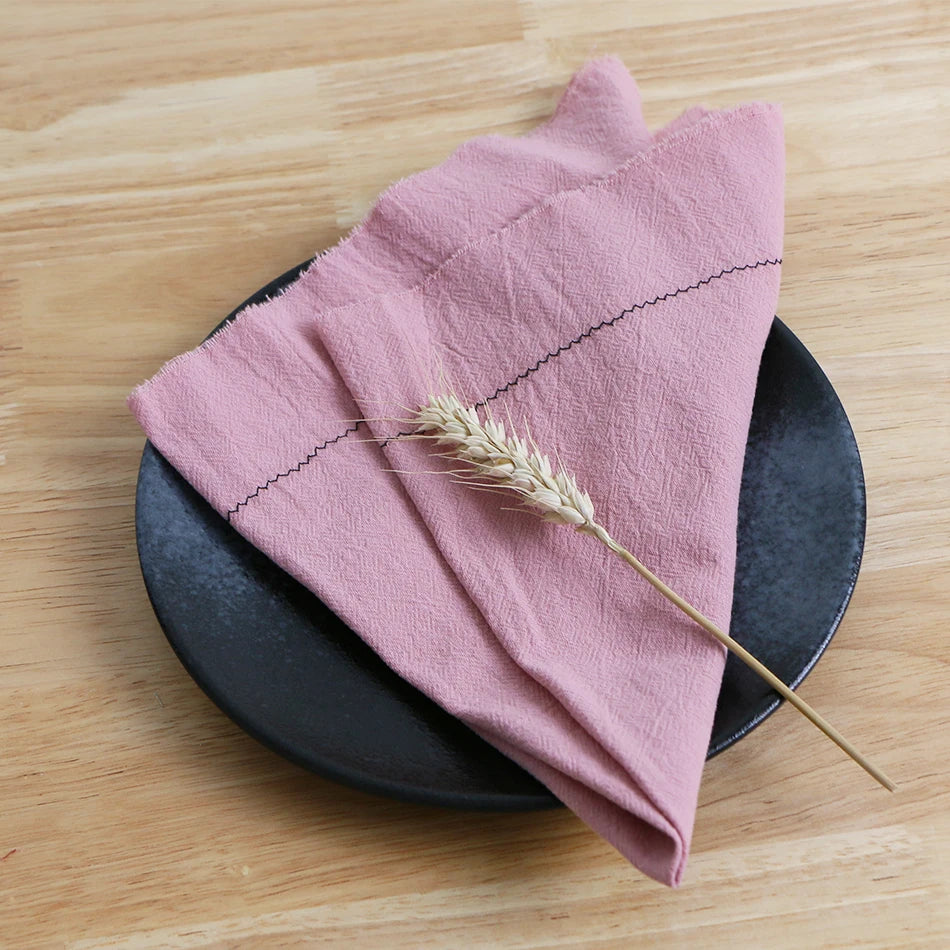 Une serviette de table rose pliée avec une épie de blé posée dessus, sur une assiette noire reposant sur une surface en bois.