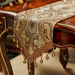 Cette image montre un magnifique chemin de table orné de motifs paisley dorés et de franges pendantes, reposant sur une élégante table en bois.