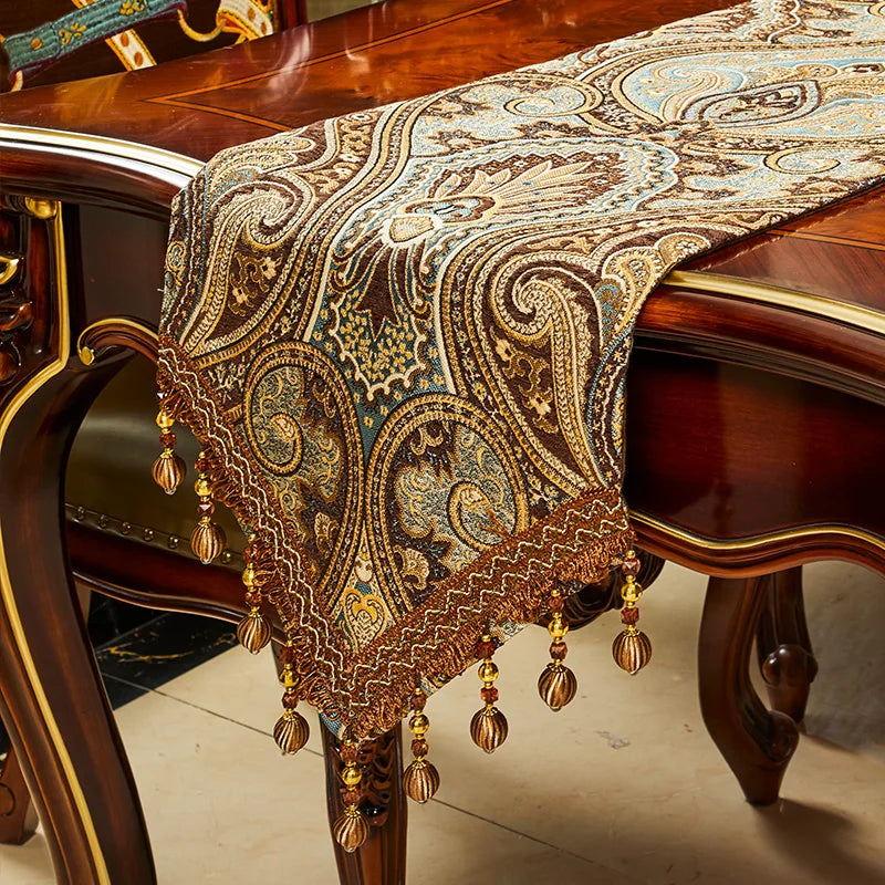 Cette image montre un magnifique chemin de table orné de motifs paisley dorés et de franges pendantes, reposant sur une élégante table en bois.
