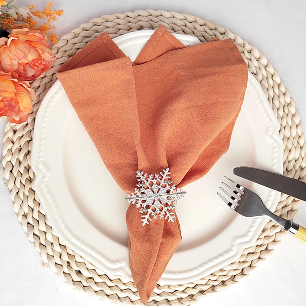 Une assiette blanche avec un napperon tressé et des serviettes orange brique, ornées d'un beau jonc en métal décoré de flocons de neige.