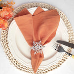 Une assiette blanche avec un napperon tressé et des serviettes orange brique, ornées d'un beau jonc en métal décoré de flocons de neige.
