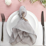 Une belle nappe en lin gris enroulée sur une assiette blanche, entourée de fleurs blanches sur un fond rose pastel.