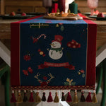 Une nappe de table décorée d'un bonhomme de neige souriant et d'ornements de Noël sur un fond bleu profond, invitant à la joie et à la célébration des fêtes.