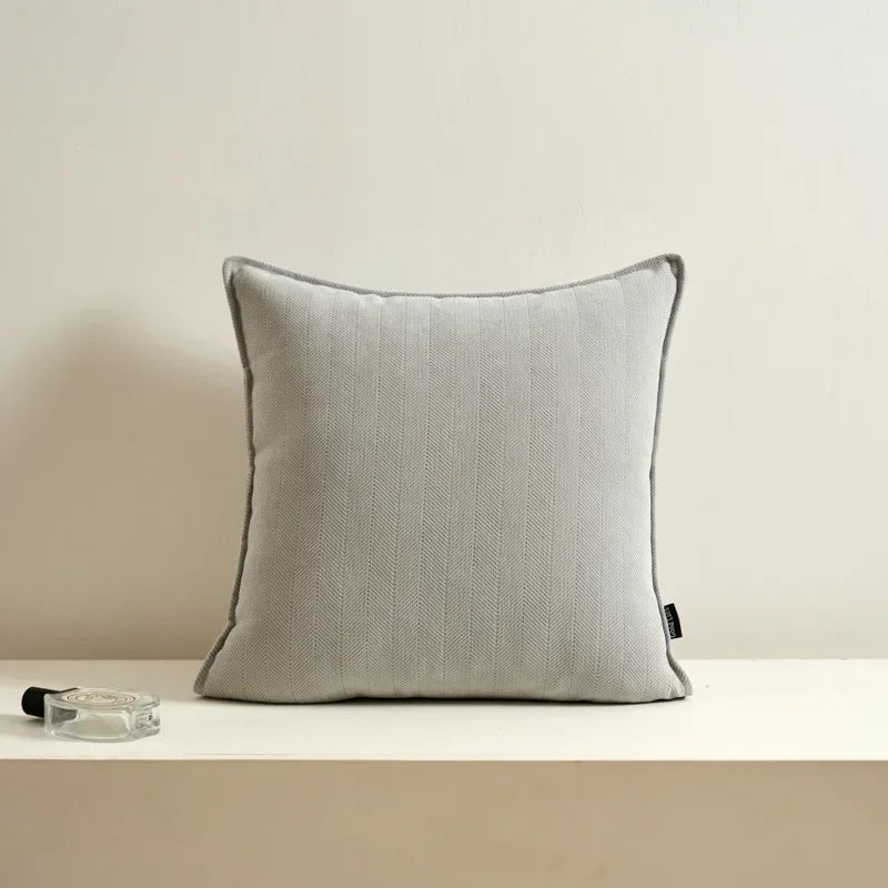 Coussin décoratif gris clair posé sur un meuble blanc avec un flacon en verre à côté.