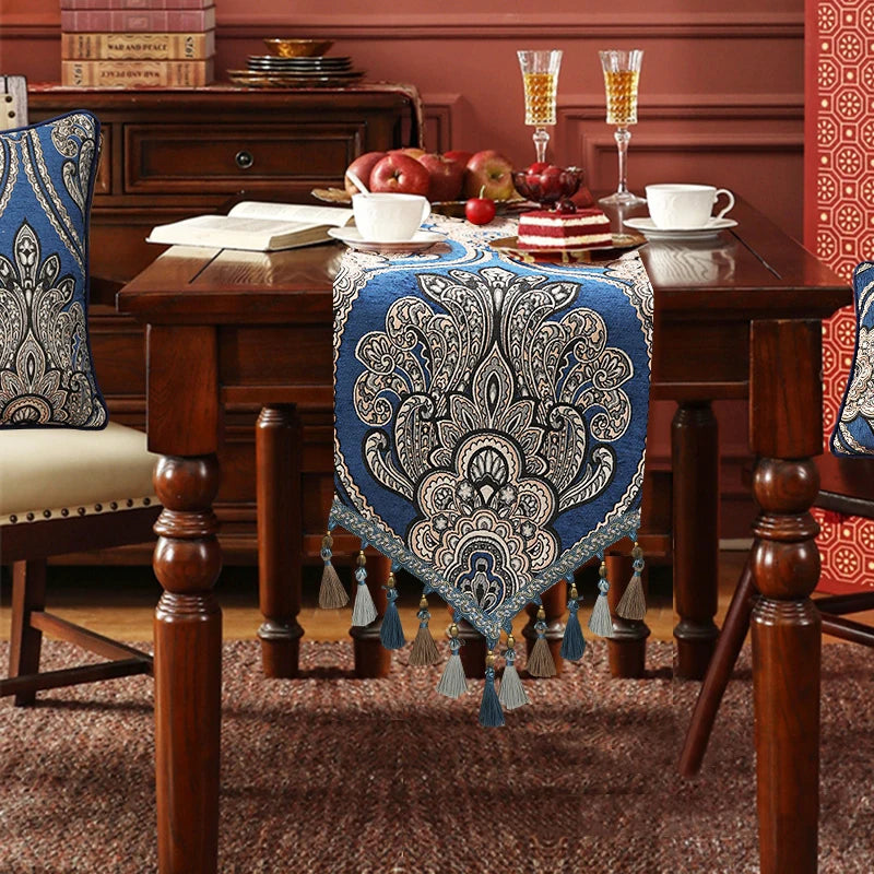 Une élégante nappe de table aux motifs floraux bleus et beige est posée sur un buffet en bois richement décoré, sur lequel se trouve un plateau avec des fruits et des desserts.