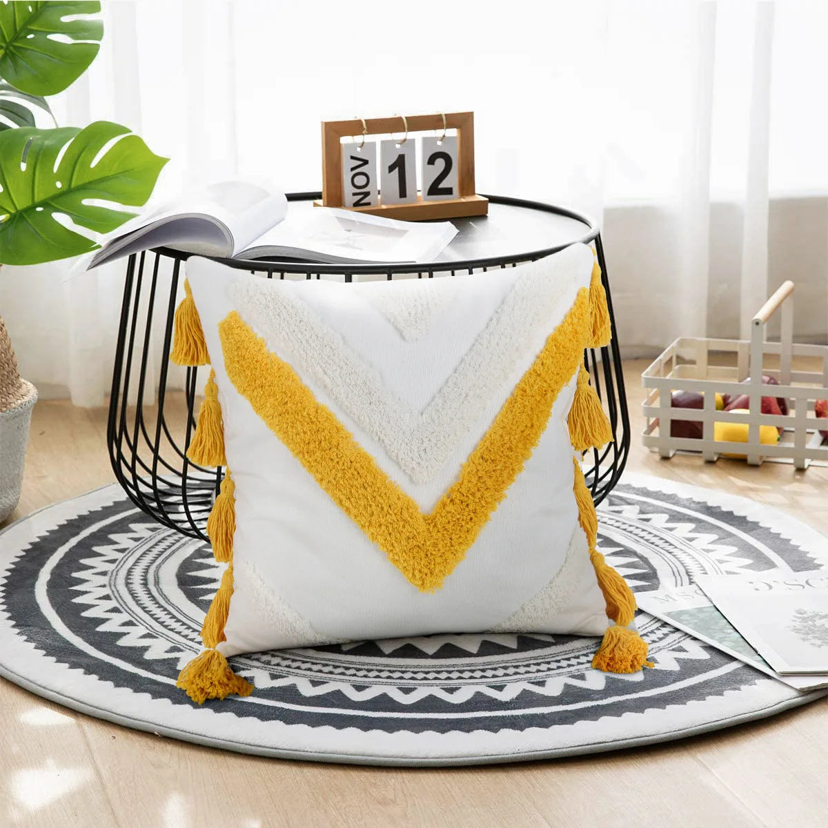 Coussin décoratif en tissu blanc et jaune avec des franges et un motif géométrique, posé sur un tapis rond motif tribal, avec un calendrier en bois à côté.