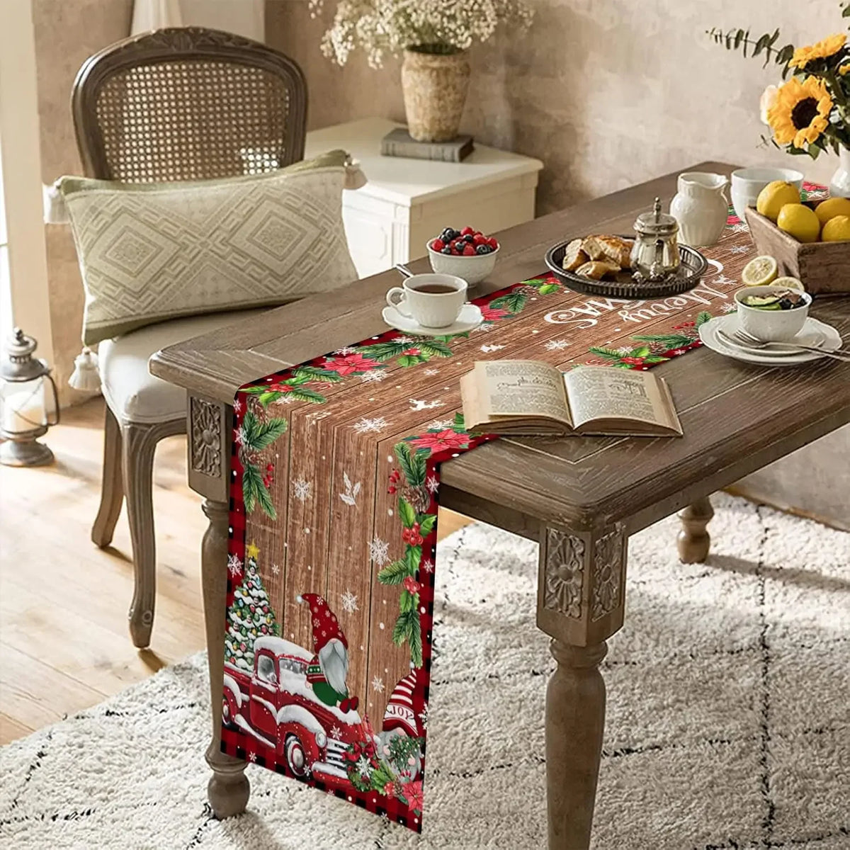 Une table de salle à manger joliment décorée dans un style rustique pour les fêtes de Noël, avec un chemin de table en tissu imprimé de motifs de Noël et de camions rouges vintage, un livre ouvert posé dessus, et des éléments de décoration comme des fleurs et des fruits.