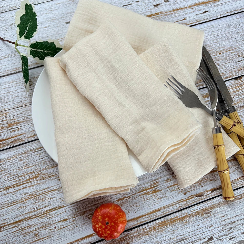 Sur une table en bois vieilli, des serviettes de table beige en lin sont posées avec une fourchette et un couteau. Une pomme rouge est également présente sur la table.