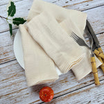 Sur une table en bois vieilli, des serviettes de table beige en lin sont posées avec une fourchette et un couteau. Une pomme rouge est également présente sur la table.