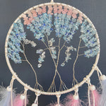 Une couronne de rêve sculptée en fil de cuivre ornée de pierres précieuses de différentes teintes de bleu et de rose, suspendue dans un cadre en forme de cercle.