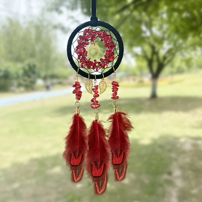 Un capteur de rêves décoratif suspendu, ornée de pierres rouges brillantes et de plumes rouges de style bohème, sur un fond flou de verdure.