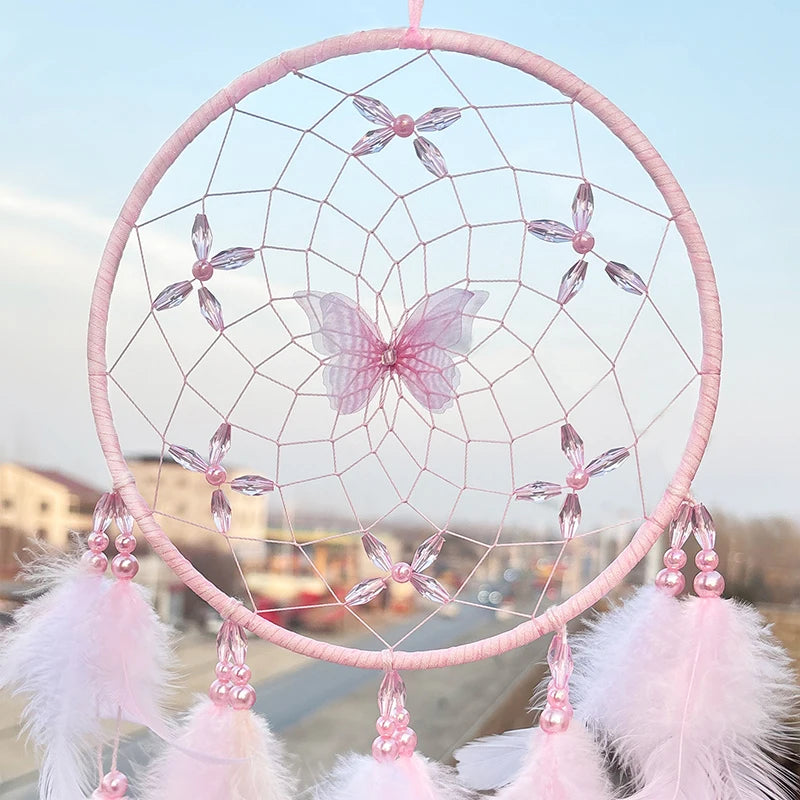 L'image montre un attrapeur de rêves rose pastel avec des perles, des plumes et une fleur décorative au centre. Cet attrapeur de rêves est suspendu dans un cadre circulaire et est composé d'un motif en nid d'abeille.