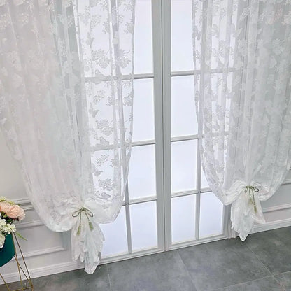Détail du voilage papillon blanc installé dans une pièce aux murs moulurés, avec un cache-pot bleu et fleurs roses.