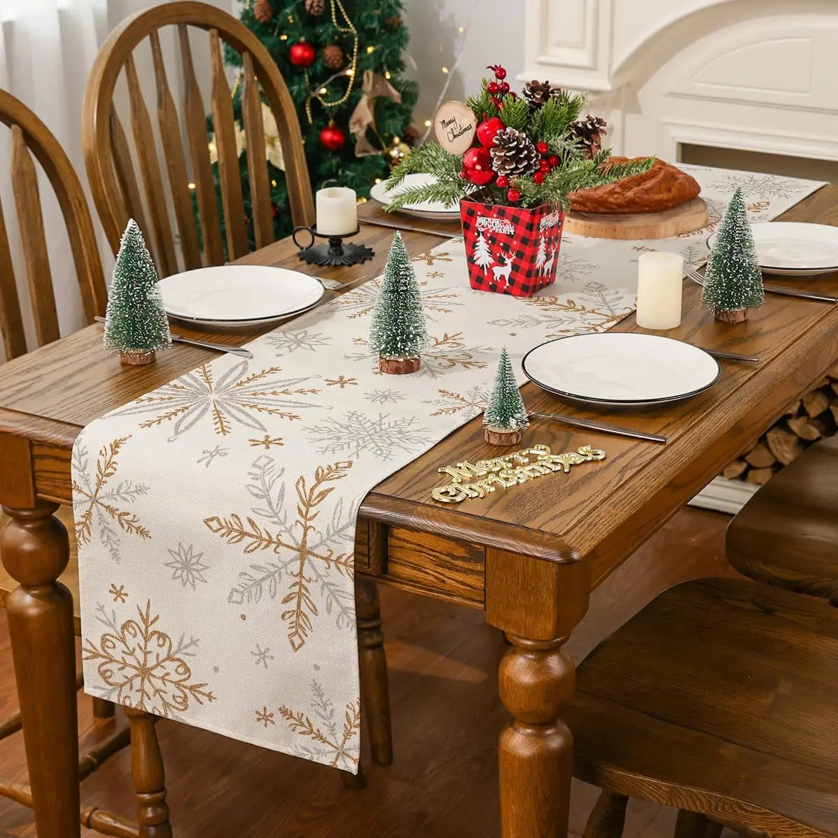 Une table de salle à manger joliment décorée pour les fêtes de Noël, avec une nappe en tissu beige aux motifs de flocons de neige, des assiettes blanches, des bougies et de petits arbres de Noël en décorations.