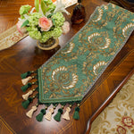 Un chemin de table vert et doré orné de motifs floraux et de pompons élégants, posé sur un meuble en bois sombre, à côté d'un vase contenant un bouquet de fleurs fraîches.