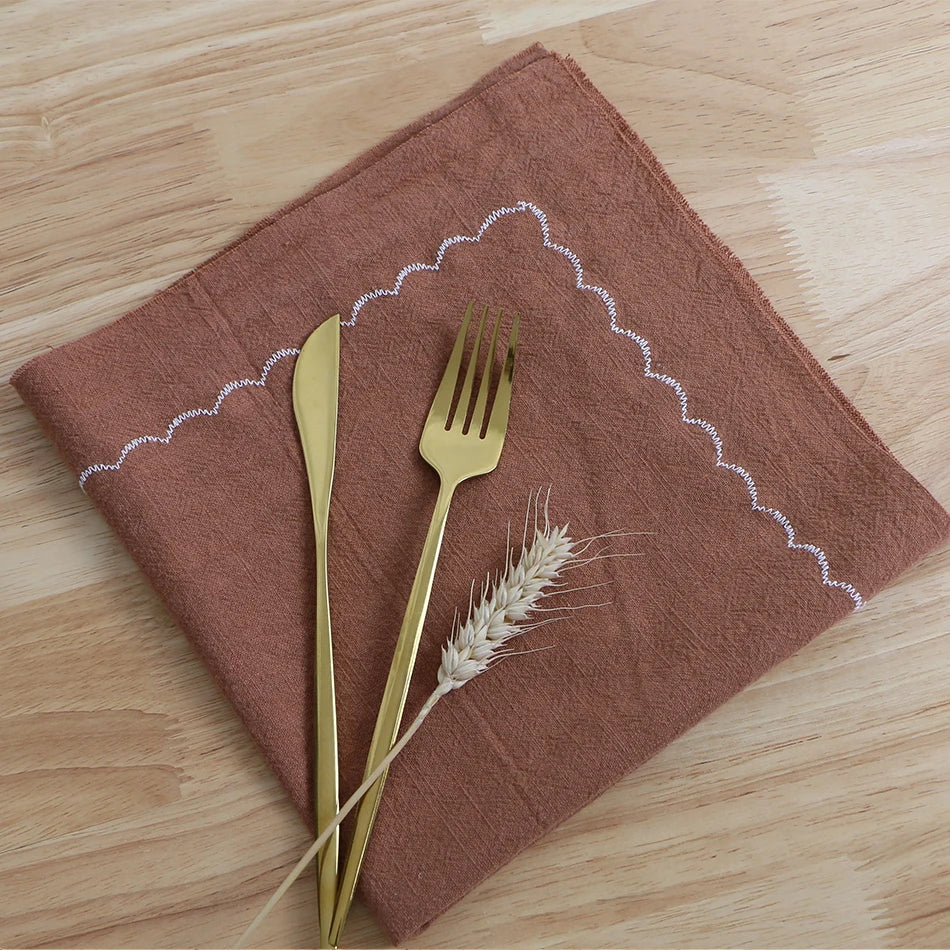 Une serviette en tissu marron, une fourchette et un couteau en métal doré, accompagnés d'épis de blé, créent une composition élégante et naturelle sur un fond en bois clair.