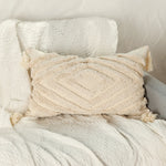 Cette image montre un coussin décoratif moelleux et texturé avec des motifs géométriques et des franges sur un tissu blanc crème, créant une ambiance douce et relaxante dans un intérieur.