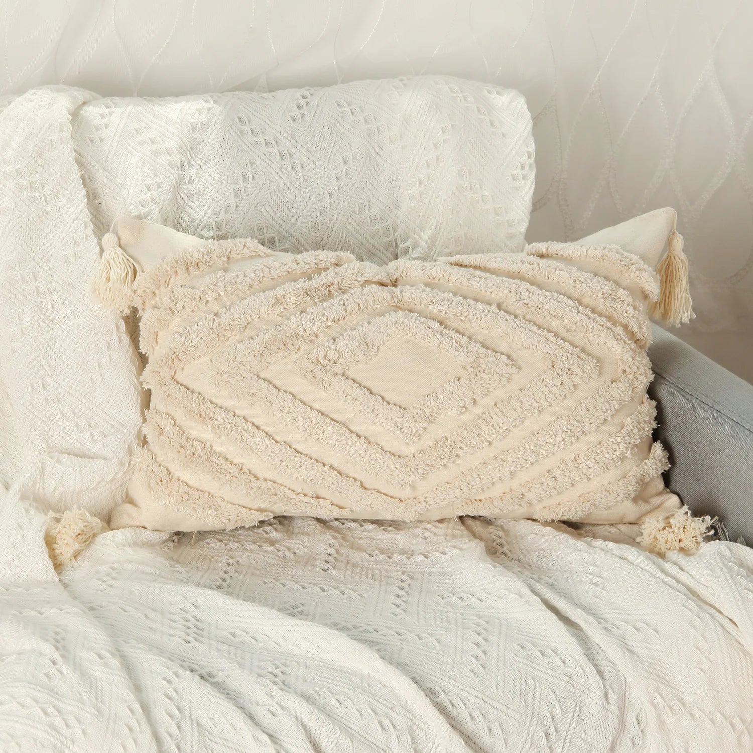 Cette image montre un coussin décoratif moelleux et texturé avec des motifs géométriques et des franges sur un tissu blanc crème, créant une ambiance douce et relaxante dans un intérieur.