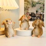 Trois petits lapins en peluche assis sur un support blanc, avec une scène estivale floue en arrière-plan.