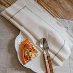 Assiette blanche avec une part de pizza garnie, accompagnée d'une fourchette et d'un couteau en bois sur une serviette en lin blanc.