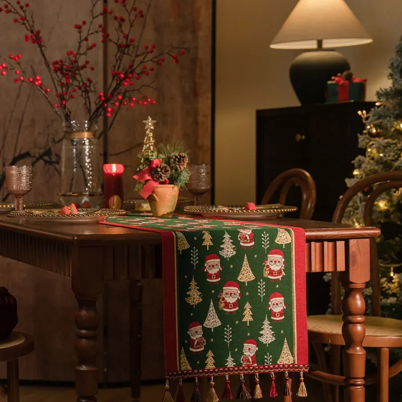 L'image montre une table de salle à manger décorée dans un style festif pour les fêtes de fin d'année, avec une nappe verte ornée de motifs de sapins et de pères Noël, des branches de baies rouges dans un vase, et un petit sapin de Noël décoré posé sur la table.