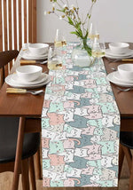 Table élégamment dressée avec une nappe colorée aux motifs de chats, des verres, des assiettes blanches et des fleurs dans un vase, créant une ambiance chaleureuse et accueillante.