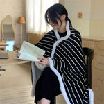 Une jeune femme portant un poncho rayé noir et blanc est assise à un bureau et lit un livre, dans un environnement intérieur confortable.