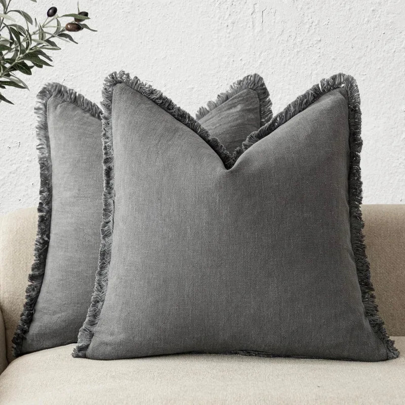 Coussin décoratif gris avec bords effilochés, appuyé sur un mur blanc avec une plante verte en arrière-plan.