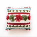 Coussin décoratif aux motifs géométriques et pompons rouges et verts sur fond blanc.