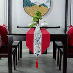 Cette image montre une table à manger élégante avec un motif floral et oriental sur un chemin de table rouge, complétée par un bonsaï vert et un cadre mural décoré de montagnes dans des tons bleutés.