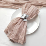 Un élégant set de table composé d'une serviette en tissu rose pâle élégamment nouée autour d'une assiette blanche, avec un élément décoratif sous forme de feuillage argenté.