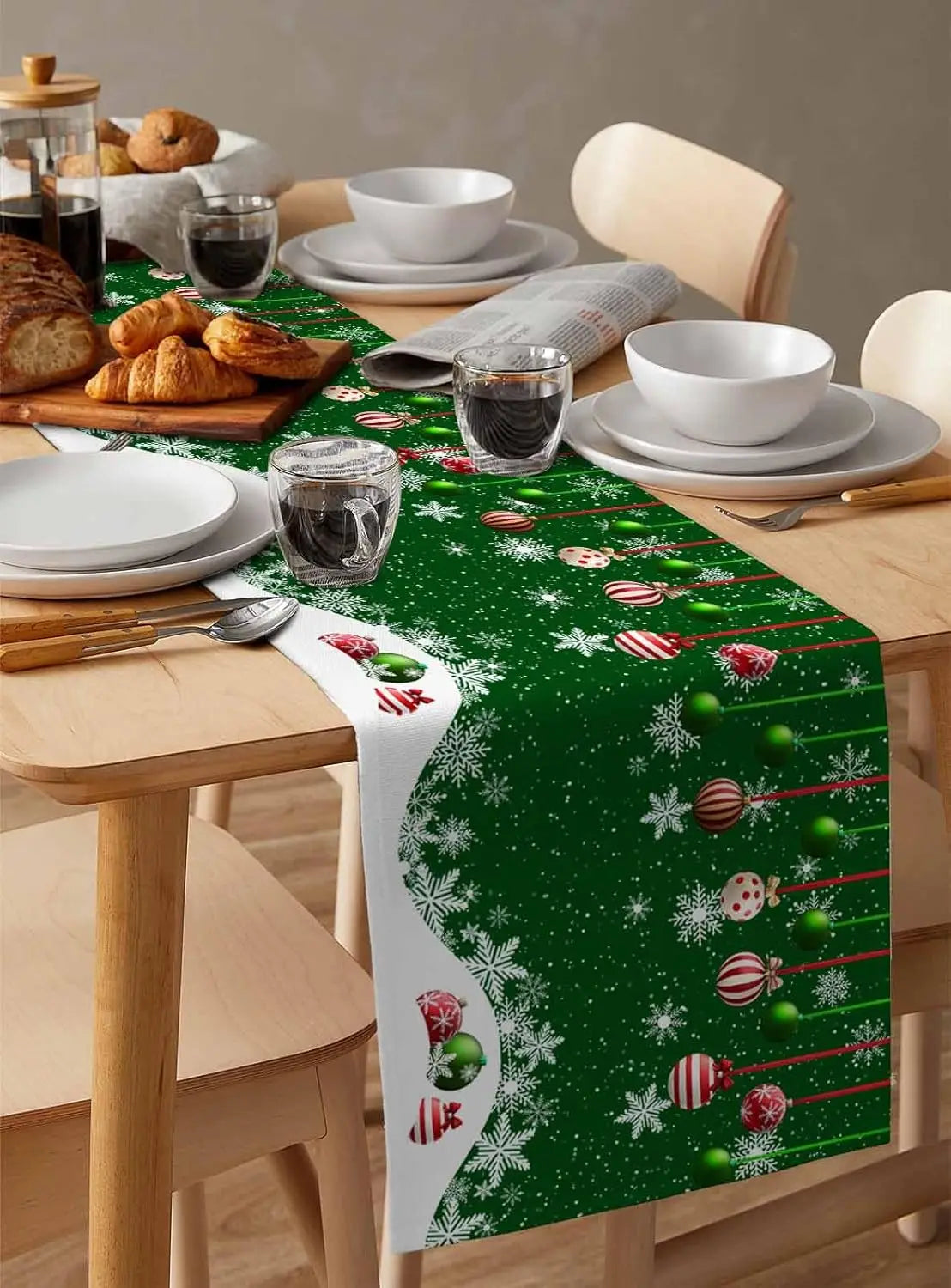 Une table de cuisine joliment décorée pour les fêtes, avec des plats blancs, des pâtisseries et un chemin de table vert festif orné de flocons de neige et de boules de Noël colorées.