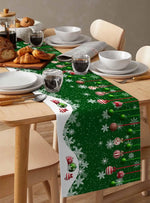 Une table de cuisine joliment décorée pour les fêtes, avec des plats blancs, des pâtisseries et un chemin de table vert festif orné de flocons de neige et de boules de Noël colorées.