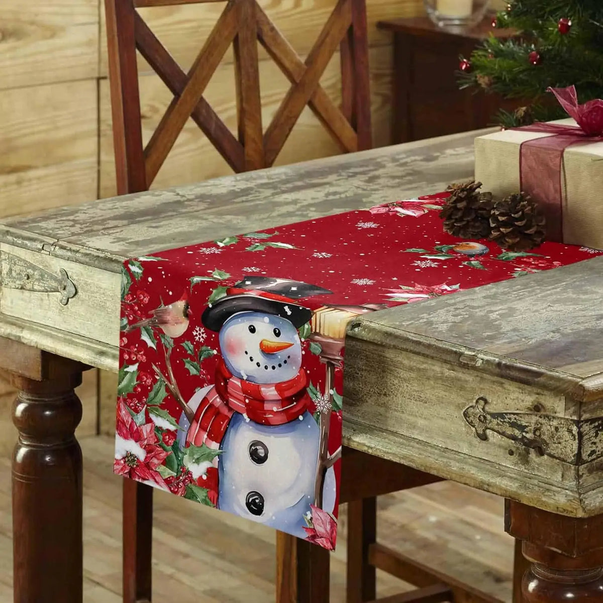 Nappe de Noël rouge ornée d'un joli bonhomme de neige assis sur une antique table en bois, avec des guirlandes et des pins sur fond de chaises en bois rustiques.