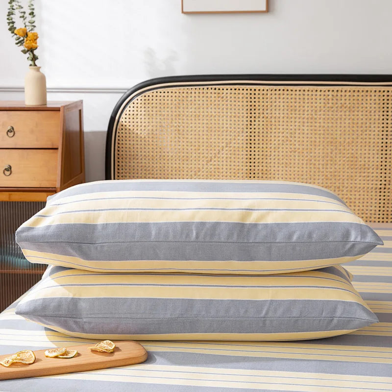 Deux oreillers confortables avec une housse rayée de couleurs neutres et douces, posés sur un lit avec une tête de lit en rotin, contre un mur blanc avec une petite vase de fleurs décorative sur un meuble de rangement en bois.