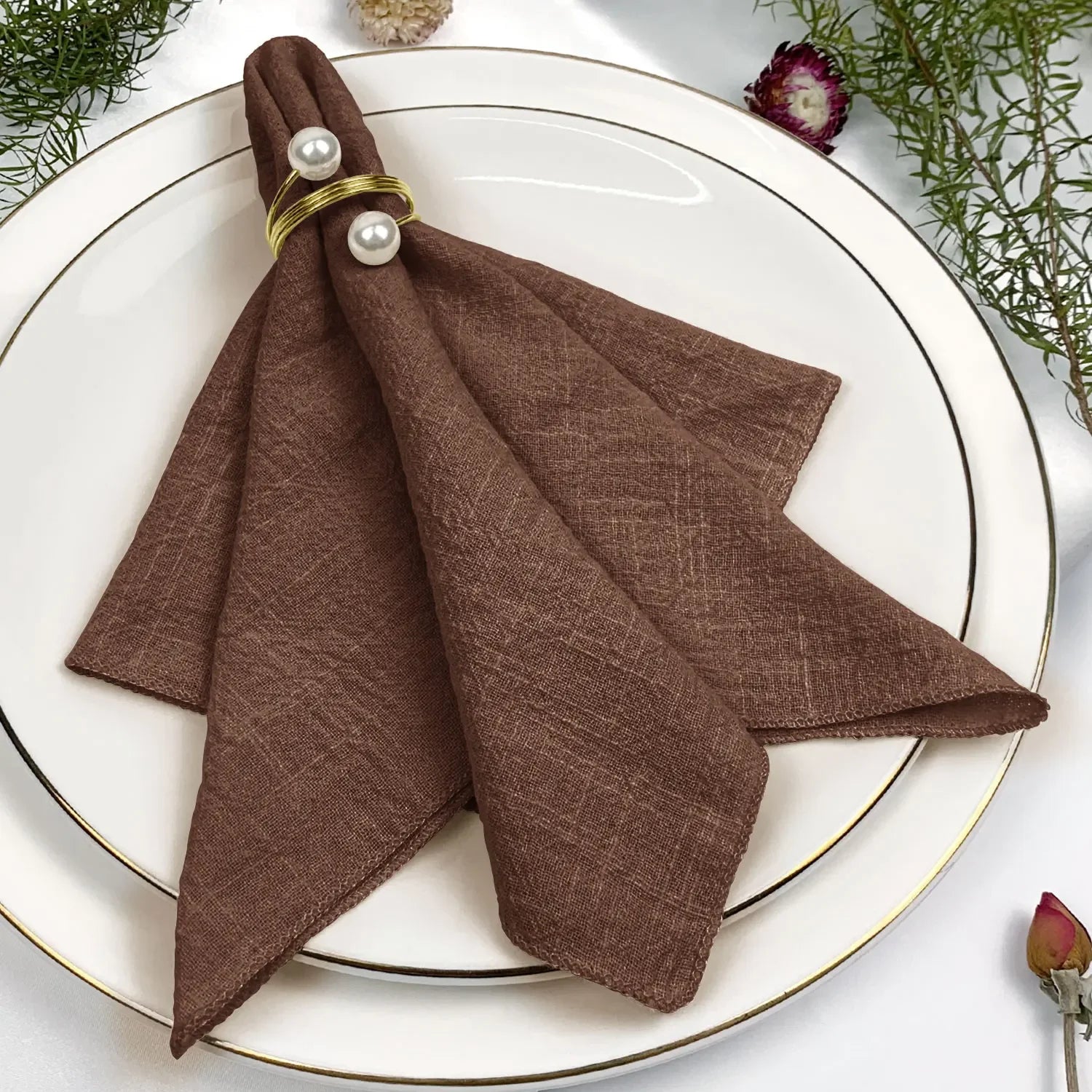 Serviettes de table luxueuses en tissu brun foncé décorées de perles, présentées sur une assiette blanche entourée de fleurs colorées.