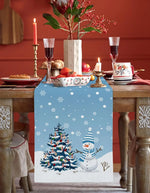 Une table de Noël joliment décorée avec un centre de table festif, des verres à vin et une bougie allumée, rehaussée par un chemin de table décoré d'un motif de flocons de neige et d'un bonhomme de neige.
