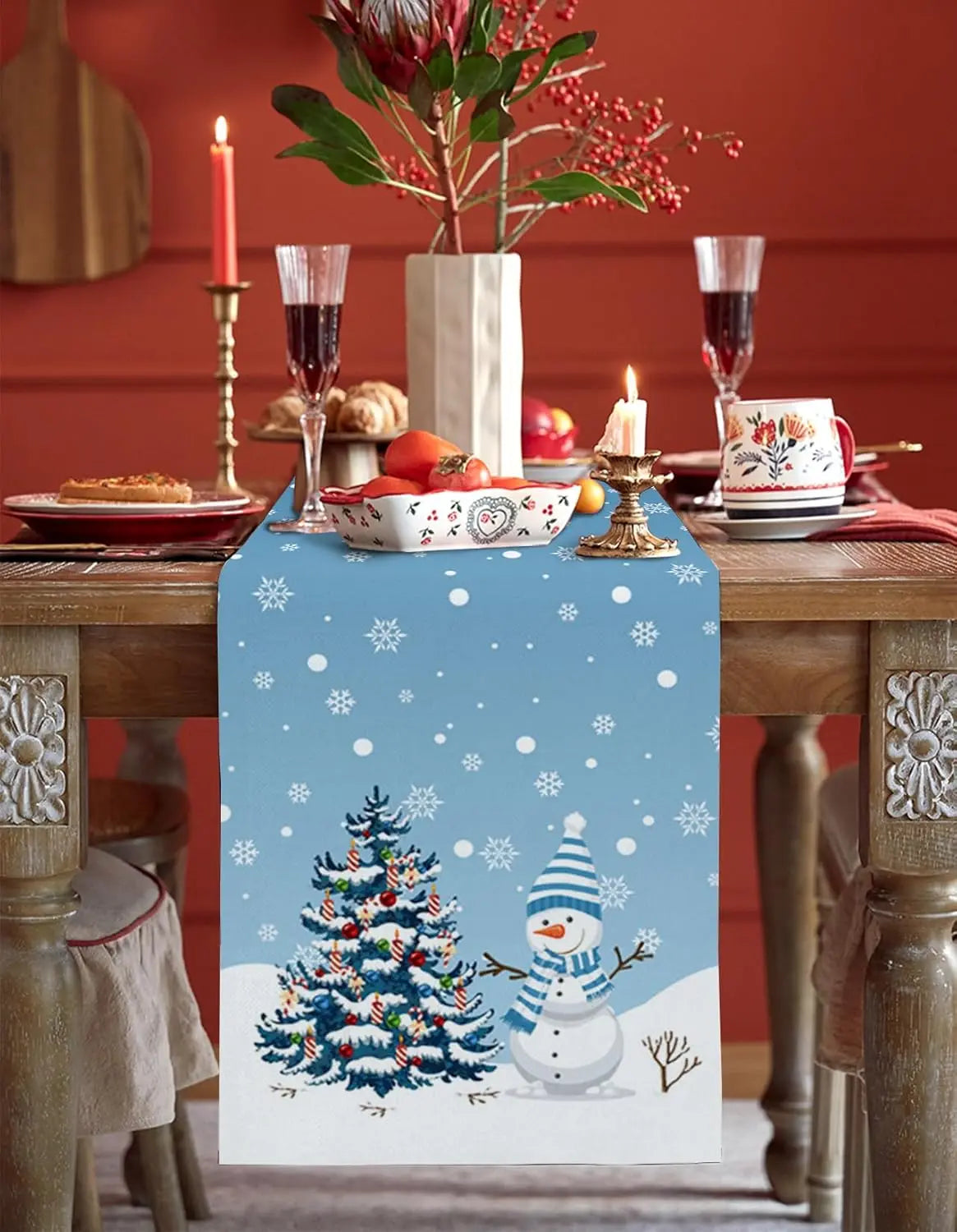 Une table de Noël joliment décorée avec un centre de table festif, des verres à vin et une bougie allumée, rehaussée par un chemin de table décoré d'un motif de flocons de neige et d'un bonhomme de neige.