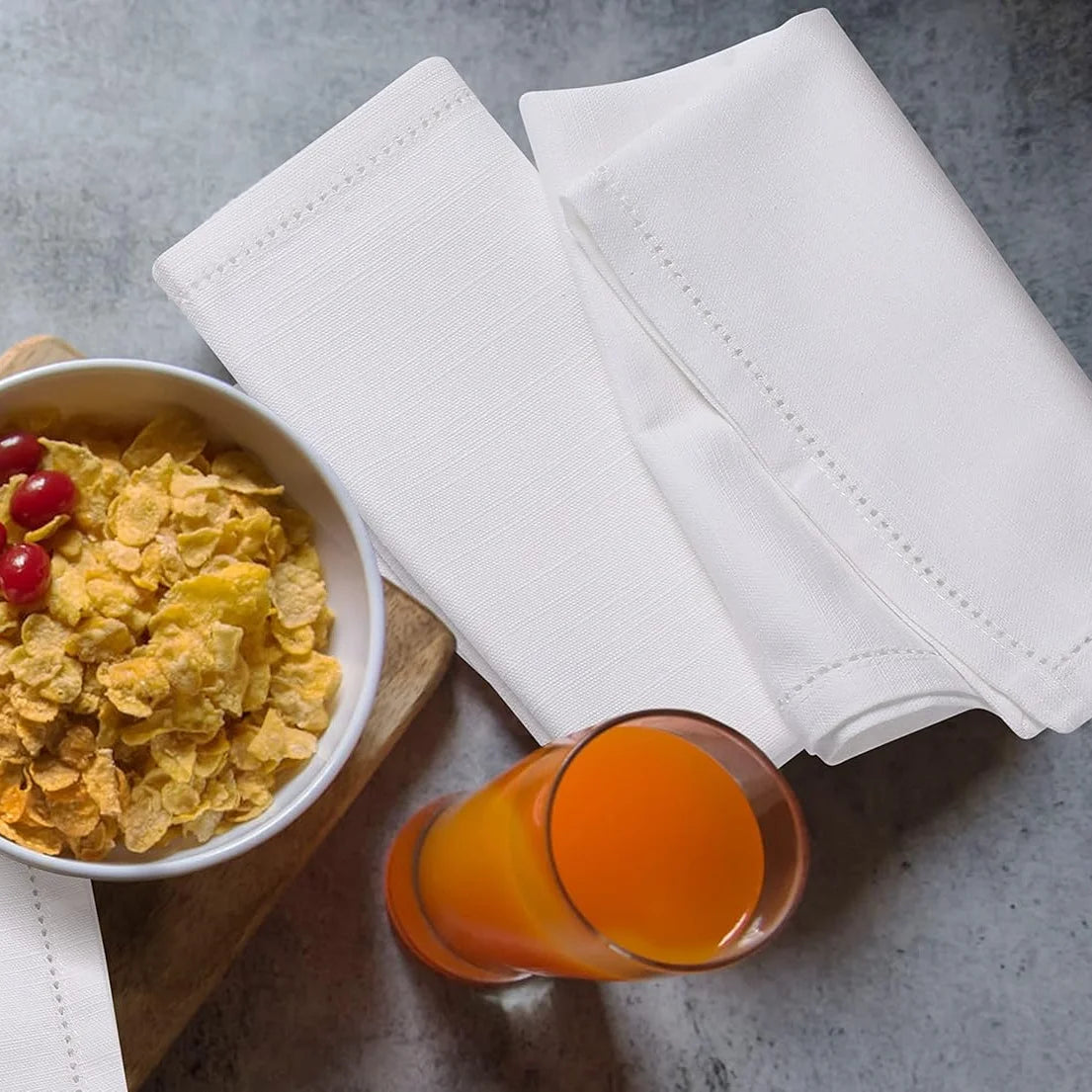Un bol de corn flakes accompagné de serviettes blanches et d'un verre de jus d'orange sur un plan de travail en béton.