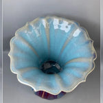 Une élégante fleur céramique en bleu clair et blanc, avec un centre foncé, présentée sur un fond gris.