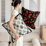 Deux coussins décoratifs aux motifs fleuris, l'un à fleurs noires sur fond blanc, l'autre à fleurs rouges vives sur fond noir, disposés près d'une fenêtre dans une pièce aux murs blancs.
