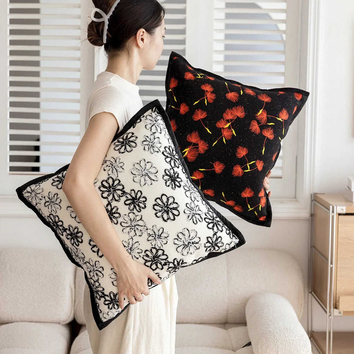 Deux coussins décoratifs aux motifs fleuris, l'un à fleurs noires sur fond blanc, l'autre à fleurs rouges vives sur fond noir, disposés près d'une fenêtre dans une pièce aux murs blancs.