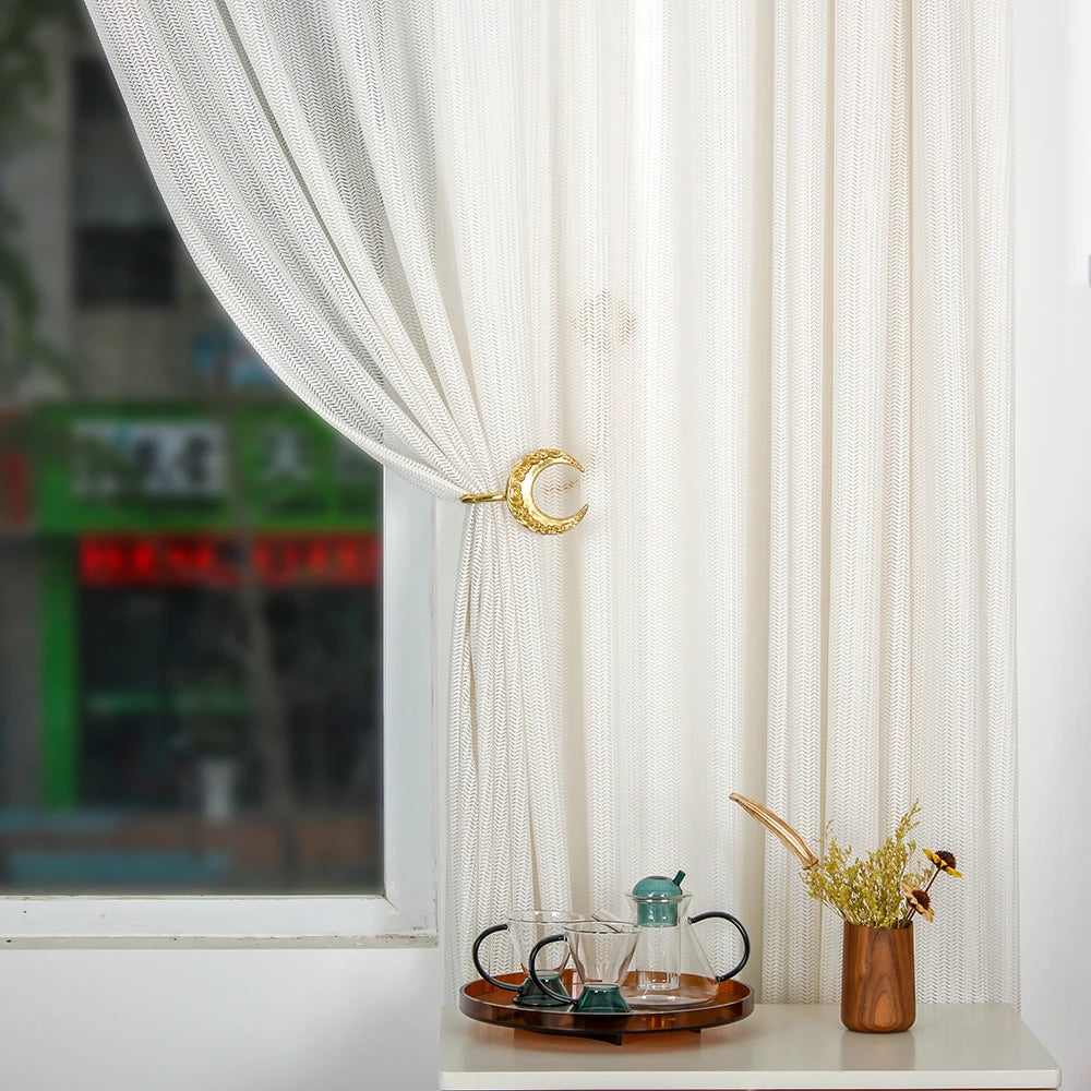 Le rideau blanc translucide avec l'accent doré en forme de lune crée une ambiance douce et paisible, complétée par le plateau en bois et les accessoires de décoration dans le coin de la pièce.
