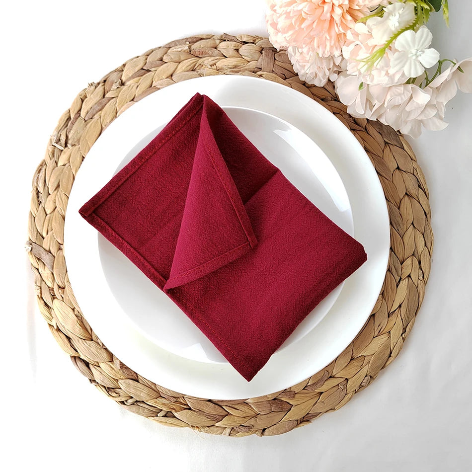 Une jolie nappe de table tressée sert de support à une assiette blanche avec une serviette en tissu rouge bordeau pliée au centre, accompagnée de fleurs colorées.