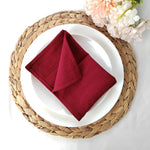 Une jolie nappe de table tressée sert de support à une assiette blanche avec une serviette en tissu rouge bordeau pliée au centre, accompagnée de fleurs colorées.