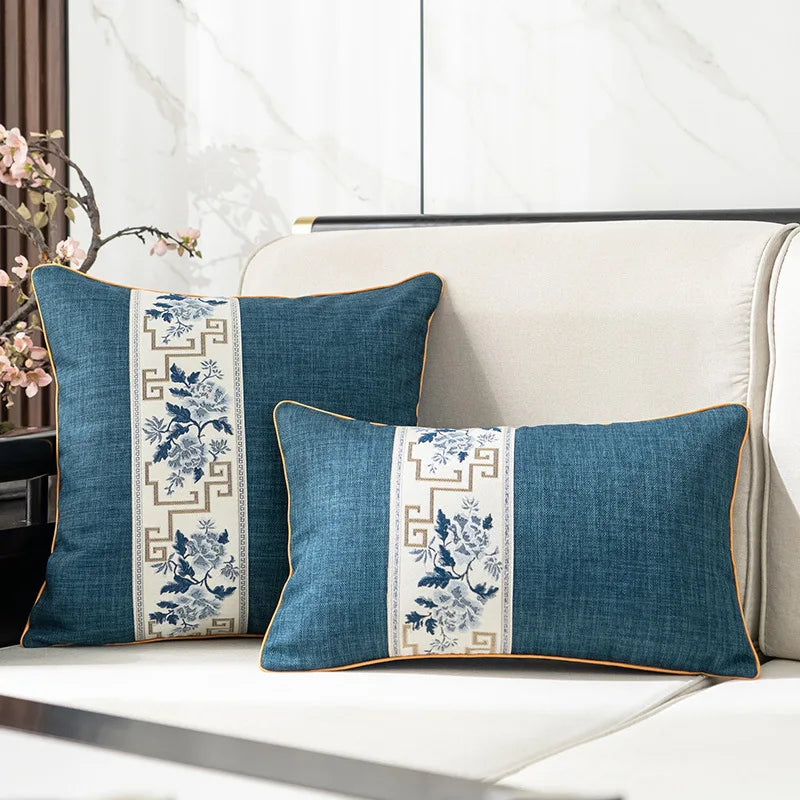 Deux coussins décoratifs bleu foncé avec des motifs floraux blancs et un encadrement doré, posés sur un canapé blanc moderne dans un intérieur lumineux avec une suspension en métal.